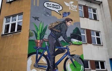 Mural przedstawia chłopaka jadącego na&nbsp;rowerze. Podczas jazdy wypowiada hasło &quot;Daj miastu detchnąć&quot;. Na bagazniku niebieskiego roweru znajduje się zielony kwiat, obok rowerzysty lecą ptaki, w&nbsp;tle szare bloki i&nbsp;zielony autobus.