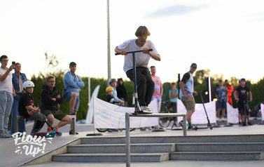 Skateplaza. Na pierwszym planie na barierce młody chłopak na hulajnodze. W tle grupa ludzi, dzieci i młodzieży obserwująca wyczyny.