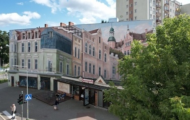 Mural na ścianie budynku przedstawiający kolorowe kamieniczki, mieszkańc&oacute;w w oknach. W parterze budynku lodziarnia. Ludzie przechadzający chodnikiem, na pierwszym planie drzewo.
