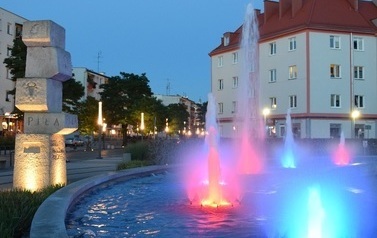 podświetlona kolorowo fontanna, w tle budynki i pomnik