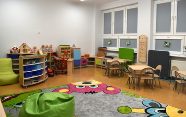 Sala przedszkolna. Na środku leży kolorowy dywan w misie, po prawej stronie okna, stoliki i krzesełka dla dzieci. Na wprost p&oacute;łki i pojemniki na zabawki, pomoce dydaktyczne. 