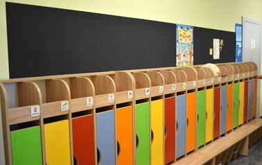Szatnia przedszkolna. Przy ścianie stoi rząd kolorowych szafek dla dzieci. Nad szafkami ściana pomalowana jest farbą magnetyczną, na kt&oacute;rej można wieszać prace dzieci.