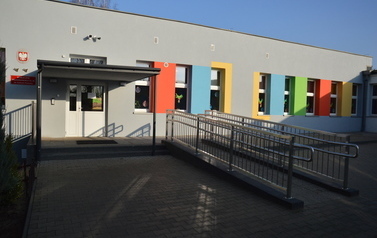 Budynek przedszkola pomalowany na szaro z kolorowymi elementami. Gł&oacute;wne wejście do budynku i podjazd dla w&oacute;zk&oacute;w.