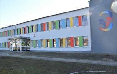 Budynek przedszkola, pomalowany na szaro z kolorowymi elementami pomiędzy oknami. Widać gł&oacute;wne wejście do budynku, po prawej stronie logo przedszkola namalowane na ścianie.