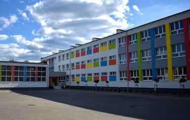 Budynek szkoły od strony boiska. Elewacja jest szara z kolorowymi elementami, część budynku to prawdopodobnie hala sportowa. W części łączącej szkołę z halą sportową znajduje się wejście do budynku.