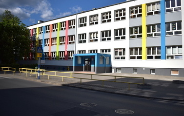 Budynek szkoły. Po lewej stronie zieleń drzew, na pierwszym planie fragment jezdni, chodnik przed szkołą i znak informujący o przejściu dla pieszych. Budynek szkoły jest szary z kolorowymi elementami. Widać gł&oacute;wne wejście do budynku.