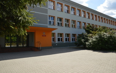 Budynek szkoły. Elewacja budynku jest po remoncie, szaro-pomarańczowa, wok&oacute;ł zieleń drzew i krzew&oacute;w. 