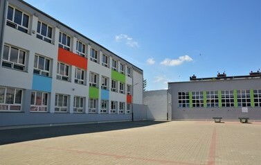 Budynek szkoły od strony boiska. Elewacja jest szara z kolorowymi elementami, część budynku to prawdopodobnie hala sportowa.