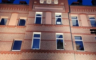 Fragment budynku z czerwonej cegły o zmroku, widać iluminacje, kt&oacute;re oświetlają gmach.