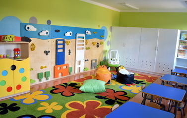 Sala w żłobku. Ściany pomalowane na zielono, w koła w innych kolorach. Po lewej stronie stoi kolorowa szafa na zabawki i ścianka wspinaczkowa. Na środku leży dywan, pufy do siedzenia, jeździk. Po prawej stronie niebieskie stoliki z krzesełkami.