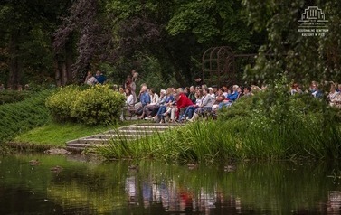 Grupa ludzi, kt&oacute;rzy siedząc na ławeczkach w parku tworzą widownię. Na pierwszym planie zbiornik wodny, dalej trzciny i krzewy, ławeczki ustawione w zielonej przestrzeni. Na dalszym planie dużo drzew i pergola. 