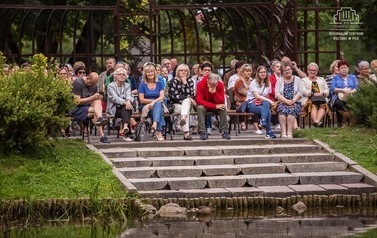 Grupa ludzi siedząca na ławeczkach w parku, za nimi pergola. Na pierwszym planie fragment zbiornika wodnego, po bokach dużo zieleni, trawa i krzewy.