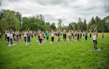 Grupa ludzi na zajęciach jogi, kt&oacute;re odbywają się na otwartej przestrzeni. Wok&oacute;ł dużo zieleni, trawa, drzewa w parku.