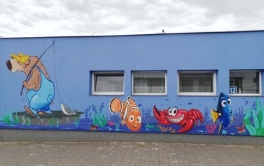 Mural na ścianie budynku przedstawia niedźwiedzia w ż&oacute;łtym kapeluszu i niebieskich ogrodniczkach, kt&oacute;ry ma na ramieniu zarzuconą wędkę. Na końcu wędki ciągnie się zahaczona ryba. po prawej stronie widać dwie ryby - błazenka i pokolca kr&oacute;lewskiego oraz kra