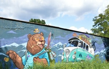 Mural przedstawia niedźwiedzia, kt&oacute;ry niesie plecak z wakacyjnymi akcesoriami - parasolem, klapkami plażowymi. Ma kapelusz i okulary przeciwsłoneczne, za nim jedzie auto tzw. og&oacute;rek, na dachu walizki, wiosła i gumowa kaczuszka. W tle muralu szczyty g&oacute;r.