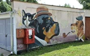 Mural na ścianie garażu przedstawia głowy dw&oacute;ch ps&oacute;w w kapeluszach, przed jednym z nich znajduje się fajka. Psy mają na głowach kapelusze, ubrane są w garnitury. Zza jednego psa wygląda kot.