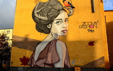 Mural na ścianie budynku przedstawia portret kobiety ubranej w zwiewną sukienkę. Dziewczyna ma związane włosy, spomiędzy nich wystaje poroże, na kt&oacute;rym siedzi ptaszek gil. Obok znajduje się napis: zoomy fashion Piła.