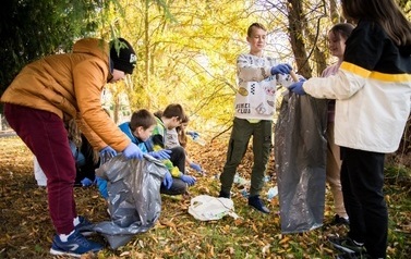 Grupa młodzieży w parku zbiera rozrzucone odpady do work&oacute;w na śmieci. Wok&oacute;ł drzewa i krzewy w jesiennych barwach.