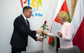 Mężczyzna wręcza kobiecie czerwoną r&oacute;że i białą teczkę.