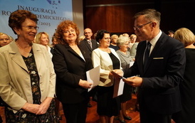 Akt immatrykulacji - mężczyzna w garniturze stoi przed grupą senior&oacute;w i wręcza dyplomy.