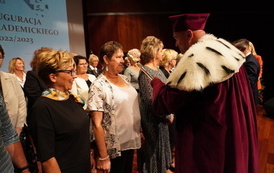 Akt immatrykulacji - rektor uczelni stoi przed grupą senior&oacute;w i dotyka berłem ramienia jednej z kobiet.