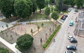 Zdjęcie z lotu ptaka, alejki parkowe. Na pierwszym planie posadzone młode drzewa, rosnąca trawa. Dalej ławeczki, posadzone młode krzewy. Na terenie także duże stare drzewa dające cień. Po prawej stronie ulica, parking a na nim auta osobowe.