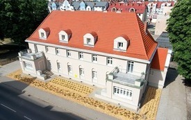 Zdjęcie z lotu ptaka. Piękna kamienica z dwoma tarasami, czerwonym dachem. Przed budynkiem nowe nasadzenia zieleni. W tle budynki mieszkalne, zieleń drzew.