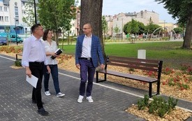 Dw&oacute;ch mężczyzn i kobieta idący ścieżką pieszą, po prawej stronie ławki, wok&oacute;ł świeże nasadzenia - młode drzewa, czerwone r&oacute;że, w oddali plac zabaw i zabudowania.
