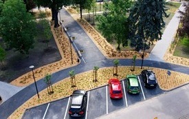 Chodniki, świeże nasadzenia roślin - młode drzewa, czerwone r&oacute;że i krzewy. W parku duże, stare drzewa r&oacute;żnego rodzaju, lampy, ławki i lampy. Na pierwszym planie parking, auta osobowe.