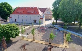Wyremontowana kamienica, szara elewacja i czerwony dach. Na pierwszym planie plac zabaw i urządzenia zabawowe, świeże nasadzenia zieleni, wysokie drzewa.