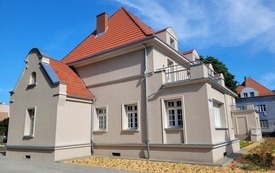 Kamienica po remoncie, szara elewacja, czerwony dach. Przed budynkiem nasadzenia zieleni, małe rośliny okrywowe.