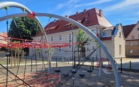 Plac zabaw, urządzenia zabawowe - siatki do wspinaczki, podłoże z kamyk&oacute;w. W tle wyremontowana kamienica, po lewej stronie duże drzewo, obok młode nasadzenia.