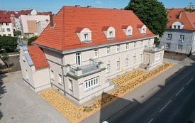 Wyremontowana kamienica z dwoma tarasami na piętrze, szara elewacja, czerwony dach. Wok&oacute;ł budynku młode nasadzenia, czerwone r&oacute;że i krzewy. W tle inne zabudowania, duże drzewo.