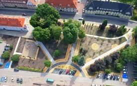 Zdjęcie z lotu ptaka. Zabudowania wielorodzinne wzdłuż drogi, obok park kieszonkowy, parkingi i plac zabaw. 