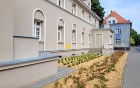 Wyremontowana kamienica z dwoma tarasami na piętrze, szara elewacja, czerwony dach. Wok&oacute;ł budynku młode nasadzenia, czerwone r&oacute;że i krzewy. W tle inna kamienica, duże drzewo.