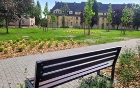 Park kieszonkowy, na pierwszym planie ławka i alejka. W tle widać zabudowę wielorodzinną. Trwa, drzewa, młode krzewy.
