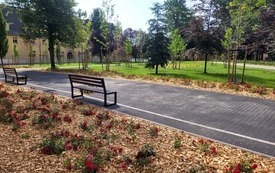 Park kieszonkowy, na pierwszym planie ławka i alejka. W tle widać zabudowę wielorodzinną. Trwa, drzewa, młode krzewy.