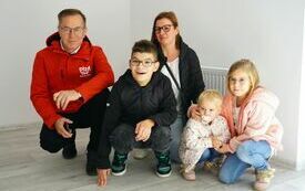 Mężczyzna, kobieta i troje dzieci kucają pozując do zdjęcia w mieszkaniu. Mężczyzn ma kurtkę z logotypem Miasto Piła, wszyscy się uśmiechają.