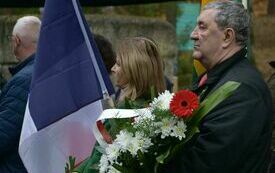 Na pierwszym planie mężczyzna trzyma wiązankę kwiat&oacute;w z biało-czerwonymi wstążkami, w tle widać flagi, ludzi i drzewa