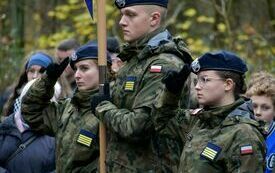 Zdjęcie przedstawia troje uczni&oacute;w klas wojskowych w mundurach stojących ze sztandarem, w tle ludzie.