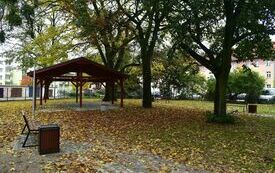 Ogr&oacute;d jesienią. Wiata piniowa, ławka i kosz na śmieci, w tle wysokie drzewa, kolorowe liście na chodniku