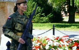Kobieta stojąca w pełnym umundurowaniu w warcie honorowej przed pomnikiem, pod kt&oacute;rym leżą wiązanki biało-czerwonych kwiat&oacute;w.