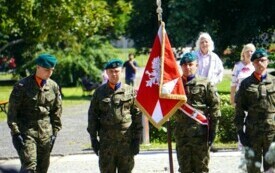 Sztandar wojska polskiego stojący w plenerze, w tle zieleń skweru, grupa ludzi.