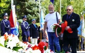 Dwoje ludzi składających wiązankę biało-czerwonych kwiat&oacute;w pod pomnikiem. W tle poczty sztandarowe.