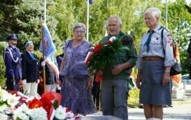 Harcerze - seniorzy składający wiązankę biało-czerwonych kwiat&oacute;w pod pomnikiem. W tle poczty sztandarowe.