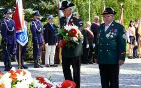Grupa ludzi w mundurach bractwa kurkowego składa wiązankę biało-czerwonych kwiat&oacute;w pod pomnikiem. W tle poczty sztandarowe.
