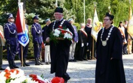 Dwoje ludzi w togach składających wiązankę biało-czerwonych kwiat&oacute;w pod pomnikiem. W tle poczty sztandarowe.