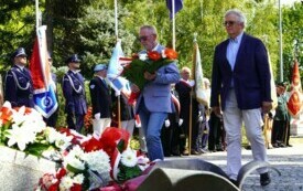 Dwoje ludzi składających wiązankę biało-czerwonych kwiat&oacute;w pod pomnikiem. W tle poczty sztandarowe.