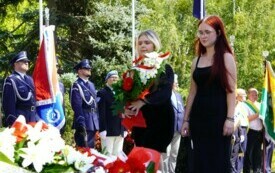 Dwie nastolatki składające wiązankę biało-czerwonych kwiat&oacute;w pod pomnikiem. W tle poczty sztandarowe.