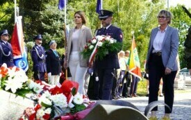 Grupa ludzi składających wiązankę biało-czerwonych kwiat&oacute;w pod pomnikiem. W tle poczty sztandarowe.